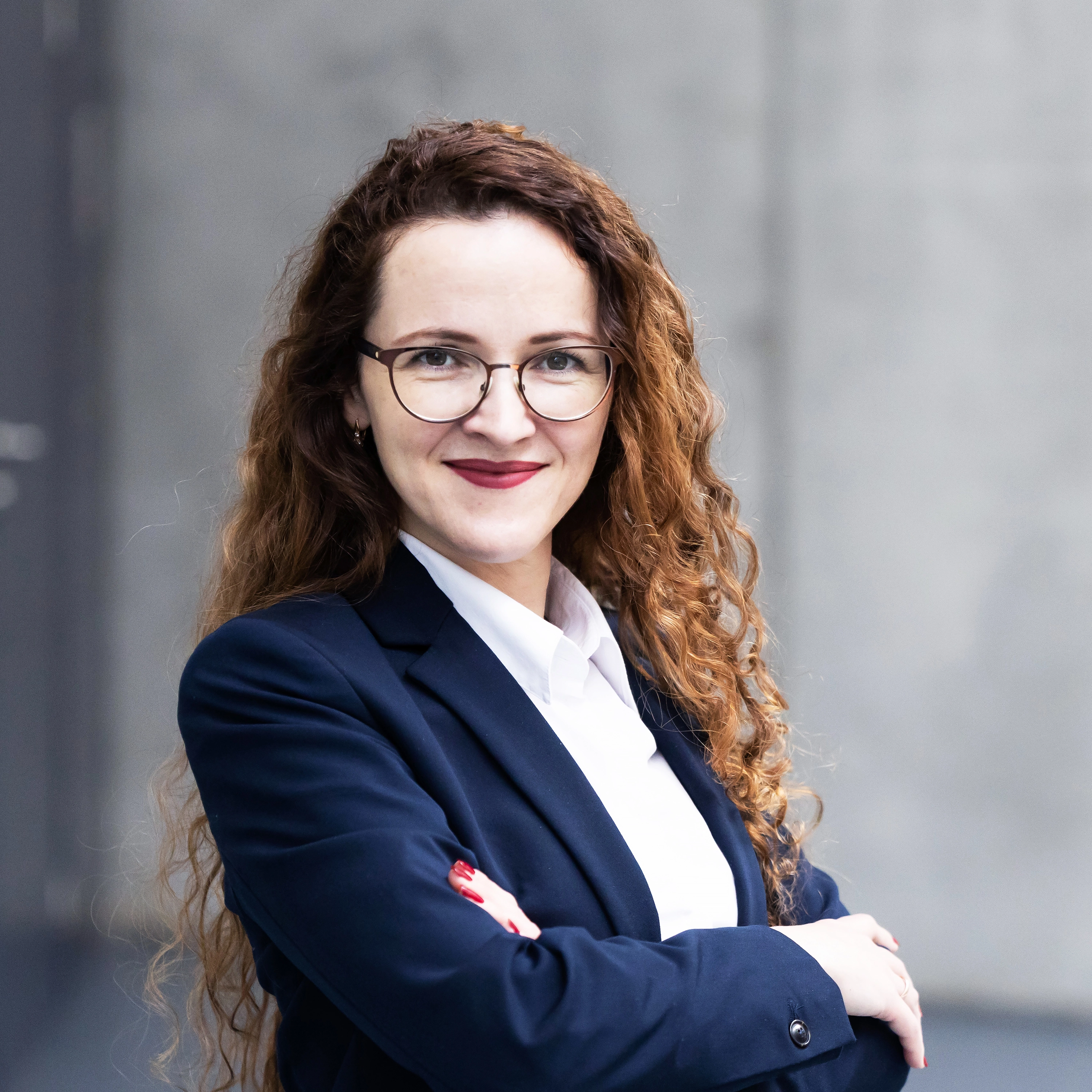 Profesionaliai atrodanti moteris su garbanotais rudais plaukais ir akiniais, vilkinti tamsiai mėlyną kostiumą ir baltus marškinius. Ji šypsosi ir atrodo pasitikinti savimi. Fonas – moderni pilkšva siena.