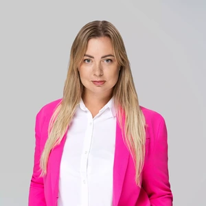 Portretinė nuotrauka – jauna moteris ilgais šviesiais plaukais, vilkinti ryškiai rožinį švarką ir baltus marškinius. Ji žiūri tiesiai į kamerą rimtu, pasitikinčiu žvilgsniu. Fonas neutralus, šviesiai pilkas.