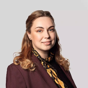 Portretinė nuotrauka – jauna moteris šviesiais, banguotais plaukais, vilkinti bordo spalvos švarku ir margu šilko šaliu. Ji žiūri į kamerą lengvai besišypsodama. Fonas neutralus, šviesiai pilkas.