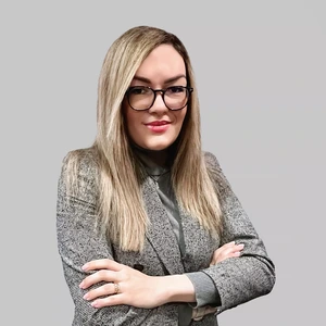 Portretinė nuotrauka – moteris ilgais šviesiais plaukais, dėvinti languotą švarką ir akinius, sukryžiavusi rankas žvelgia į kamerą šviesiame fone.