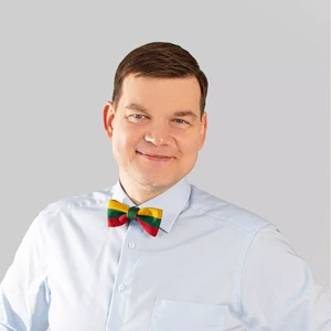 Portretinė nuotrauka – vyras trumpais rudos spalvos plaukais, vilkintis šviesiai mėlynus marškinius ir pasipuošęs peteliške su Lietuvos vėliavos spalvomis. Jis žiūri į kamerą ir šypsosi, fonas neutralus, pilkšvas.