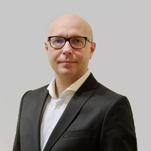 Portretinė nuotrauka – vyras be plaukų, nešiojantis juodus akinius ir auskarą, vilkintis baltus marškinius ir tamsų švarką. Jis žiūri į kamerą neutraliame, pilkame fone.
