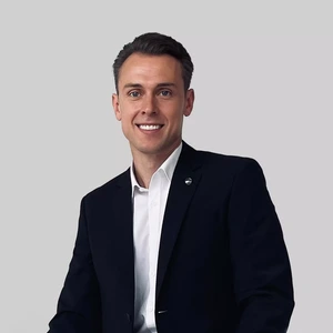 Vyresnis jaunas vyras vilki tamsų kostiumą ir baltus marškinius, šypsosi žiūrėdamas į kamerą šviesiame fone.