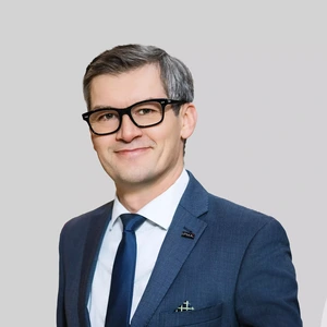 Vyresnio amžiaus vyras su akiniais ir žilstelėjusiais plaukais dėvi mėlyną kostiumą, baltus marškinius ir tamsų kaklaraištį. Jis šypsosi ir žvelgia į kamerą šviesiame fone. Ant švarko atlapo prisegtas ženkliukas su užrašu „IPMA“.