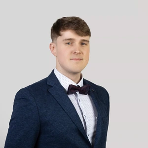 Portretinė nuotrauka – jaunas vyras trumpais šviesiai rudais plaukais, vilkintis tamsiai mėlyną kostiumą, taškuotus baltus marškinius ir bordo spalvos peteliškę. Jis žvelgia į kamerą neutraliame, pilkame fone.