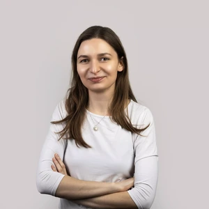 Portretinė nuotrauka – moteris rudais ilgais plaukais, vilkinti šviesius marškinėlius ir dėvinti saulės formos pakabuką, šypsosi žvelgdama į kamerą šviesiame fone, sukryžiavusi rankas.