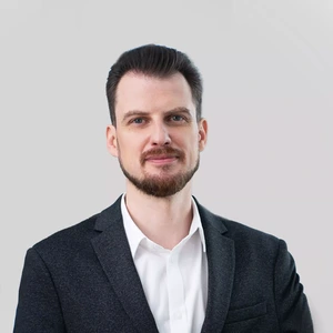 Portretinė nuotrauka – vyras tamsiais, atgal sušukuotais plaukais ir trumpa barzda, vilkintis baltus marškinius ir tamsų švarką. Jis žvelgia į kamerą švelniai šypsodamasis, fonas neutralus, pilkšvas.