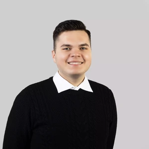 Portretinė nuotrauka – jaunas vyras trumpais tamsiais plaukais, apsirengęs baltus marškinius ir juodą megztinį su pynių raštu. Jis šypsosi ir žiūri tiesiai į kamerą, fonas – neutralus, šviesiai pilkšvas.