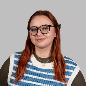 Portretinė nuotrauka – jauna moteris ilgais raudonais plaukais, dėvinti didelius juodus akinius, dryžuotą megztinį mėlynos ir baltos spalvų derinyje bei žalią palaidinę. Ji žiūri į kamerą su lengva šypsena, fonas neutralus, pilkšvas.