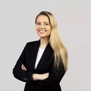Jauna moteris ilgais šviesiais plaukais, vilkinti juodu švarku ir baltu viršutiniu drabužiu, stovi sukryžiavusi rankas ir šypsosi žiūrėdama į kamerą šviesiame fone.
