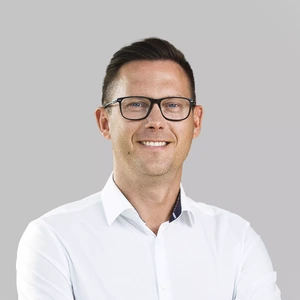Vyras trumpais tamsiais plaukais ir akiniais dėvi baltus marškinius, stovi sukryžiavęs rankas ir šypsosi žiūrėdamas į kamerą šviesiame fone.