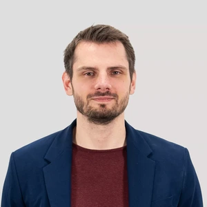 Portretinė nuotrauka – vyras trumpais rudos spalvos plaukais ir barzda, vilkintis tamsiai mėlyną švarką ir bordo spalvos marškinėlius. Jis žiūri į kamerą neutraliame, pilkame fone.