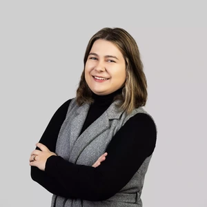 Portretinė nuotrauka – jauna moteris pusilgiais šviesiai rusvais plaukais, apsirengusi juodą golfą ir pilką liemenę. Ji šypsosi ir žvelgia į kamerą, fone – neutralus, šviesiai pilkas fonas.