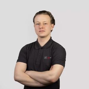 Jaunas vyras šviesiais plaukais dėvi tamsius polo marškinėlius su „ktu gifted“ logotipu, stovi sukryžiavęs rankas ir žvelgia į kamerą neutraliu veidu šviesiame fone.