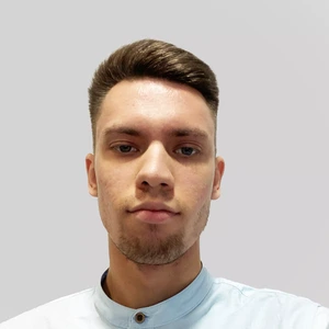 Jaunas vyras trumpais tamsiais plaukais ir vešlia barzda dėvi šviesiai mėlynus marškinius su stoveliu, žiūri į kamerą rimtu veidu šviesiame fone.