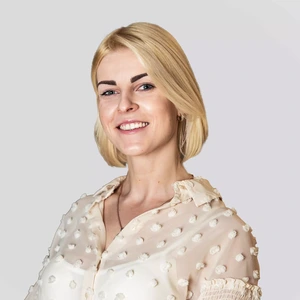 Portretinė nuotrauka – šviesiaplaukė moteris trumpais plaukais, vilkinti šviesius permatomus marškinėlius su dekoratyviais taškeliais, šypsosi žvelgdama į kamerą šviesiame fone.