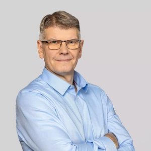 Portretinė nuotrauka – vyresnio amžiaus vyras trumpais žilais plaukais ir akiniais, vilkintis šviesiai mėlynais marškiniais. Jis žiūri į kamerą su lengva šypsena, rankos sukryžiuotos. Fonas neutralus, šviesiai pilkas.