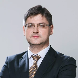 Portretinė nuotrauka – vidutinio amžiaus vyras trumpais tamsiais plaukais, su akiniais. Jis vilki tamsų kostiumą, baltus marškinius ir rudai raštuotą kaklaraištį. Žiūri į kamerą rimtu žvilgsniu. Fonas neutralus, šviesiai pilkas.