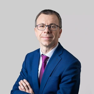 Portretinė nuotrauka – vyresnis vyras su akiniais, dėvintis tamsiai mėlyną kostiumą, šviesius marškinius su raštais ir fuksijos spalvos kaklaraištį. Jis žvelgia į kamerą su švelnia šypsena, fonas – neutralus, šviesiai pilkas.