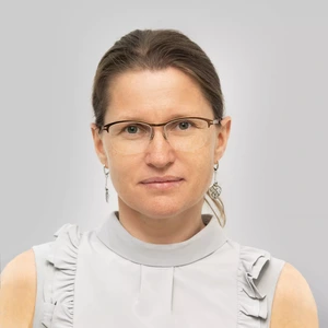 Portretinė nuotrauka – moteris su akiniais ir surištais plaukais, dėvinti šviesiai pilką palaidinę be rankovių, dekoruotą klostėmis. Ji žiūri tiesiai į kamerą, fone – šviesiai pilkas fonas