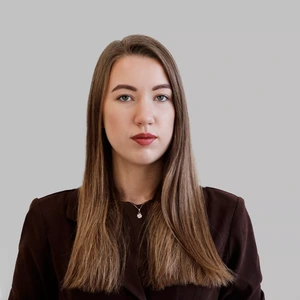 Rimta jauna moteris ilgais rudais plaukais, dėvinti tamsų švarką, pozuoja šviesiame fone.