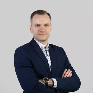 Vyras trumpais šviesiais plaukais dėvi tamsiai mėlyną kostiumą ir baltus marškinius su taškučiais, stovi sukryžiavęs rankas ir žiūri į kamerą šviesiame fone.