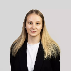 Portretinė nuotrauka – jauna moteris tiesiais šviesiais plaukais, vilkinti baltus marškinėlius ir juodą švarką. Ji žvelgia į kamerą su švelnia šypsena, fonas neutralus, pilkas.