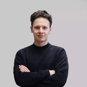 Jaunas vyras trumpais tamsiais plaukais dėvi juodą megztinį, stovi sukryžiavęs rankas ir žiūri į kamerą šviesiame fone su lengva šypsena.