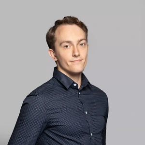 Portretinė nuotrauka – jaunas vyras trumpais tamsiais plaukais, vilkintis tamsiai mėlyną marškinius su smulkiais taškeliais. Jis žiūri į kamerą su švelnia šypsena, fonas neutralus, pilkšvas.