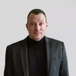 Portretinė nuotrauka – vyras trumpais tamsiais plaukais, vilkintis juodą golfą ir tamsų švarką su subtilia tekstūra. Jo žvilgsnis tiesus, rimtas. Fonas neutralus, šviesiai pilkas.