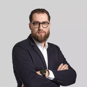 Rimtas vyras su barzda ir akiniais, dėvintis tamsų švarką ir baltus marškinius, sukryžiavęs rankas prieš šviesų foną.