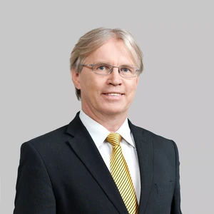 Portretinė nuotrauka – vidutinio amžiaus vyras šviesiais plaukais, dėvintis juodą kostiumą, baltus marškinius ir geltonai dryžuotą kaklaraištį. Jis žiūri į kamerą su lengva šypsena, fonas neutralus, šviesiai pilkas.