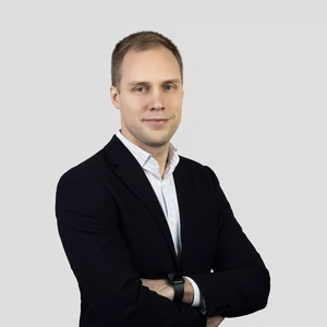 Jaunas vyras trumpais šviesiais plaukais dėvi tamsų švarką, baltus dryžuotus marškinius ir išmanųjį laikrodį. Jis stovi sukryžiavęs rankas ir žvelgia į kamerą šviesiame fone.