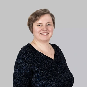 Portretinė nuotrauka – moteris trumpais šviesiais plaukais, dėvinti tamsų raštuotą viršutinį drabužį ir subtilų aukso spalvos vėrinį. Ji šypsosi žiūrėdama į kamerą. Fonas neutralus, šviesiai pilkas.