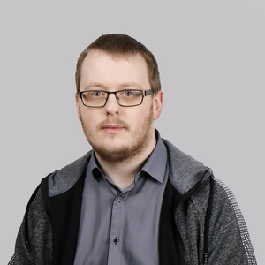 Portretinė nuotrauka – jaunas vyras trumpais plaukais ir akiniais, vilkintis pilkus marškinius ir tamsų sportinį džemperį, žvelgia tiesiai į kamerą šviesiame fone.