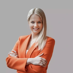 Jauna moteris ilgais šviesiais plaukais dėvi ryškiai oranžinį švarką, stovi sukryžiavusi rankas ir šypsosi žiūrėdama į kamerą pilkame fone.