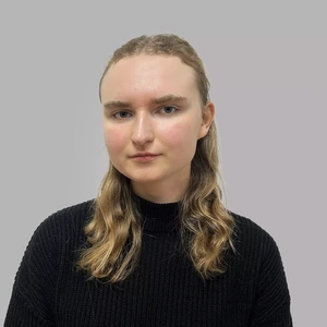 Portretinė nuotrauka – jauna moteris šviesiais banguotais plaukais, vilkinti juodą megztinį aukštu kaklu. Ji žiūri į kamerą rimtu žvilgsniu, fonas neutralus, pilkšvas.