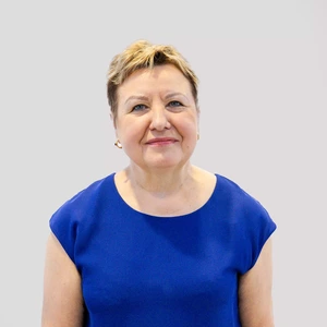 Portretinė nuotrauka – vyresnio amžiaus moteris trumpais šviesiais plaukais, vilkinti ryškiai mėlyną palaidinę, žvelgia į kamerą šviesiame fone, švelniai šypsosi.