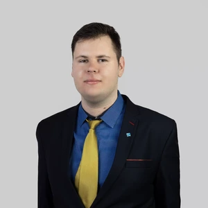 Portretinė nuotrauka – jaunas vyras trumpais plaukais, vilkintis tamsų kostiumą, mėlynus marškinius ir geltoną kaklaraištį, žvelgia į kamerą šviesiame fone. Ant švarko atlapo prisegtas ženkliukas.