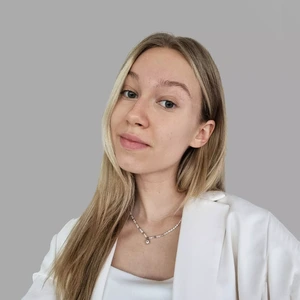 Portretinė nuotrauka – jauna moteris ilgais šviesiais plaukais, vilkinti baltą švarką ir baltą palaidinę. Ji žvelgia į kamerą šiek tiek pasukusi galvą. Fonas – neutraliai pilkas. Prie kaklo – subtili grandinėlė su pakabuku.