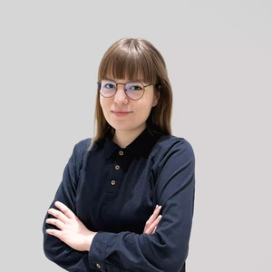 Jauna moteris su rudais plaukais ir kirpčiukais, dėvinti tamsiai mėlynus marškinėlius su sagomis ir akinius, stovi sukryžiavusi rankas prieš pilką foną.