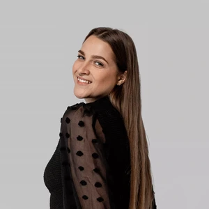 Portretinė nuotrauka – jauna moteris ilgais, tiesiais plaukais, vilkinti juodą palaidinę su permatomomis rankovėmis ir dekoratyviniais taškeliais. Ji žvelgia per petį į kamerą ir šypsosi. Fonas neutralus, šviesiai pilkas.