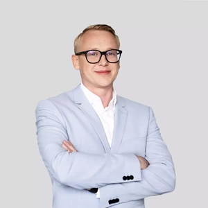Portretinė nuotrauka – vyras trumpais šviesiais plaukais, nešiojantis akinius, vilkintis šviesiai pilką kostiumą ir baltus marškinius. Jis stovi sukryžiavęs rankas, šypsosi ir žiūri į kamerą neutraliame fone.
