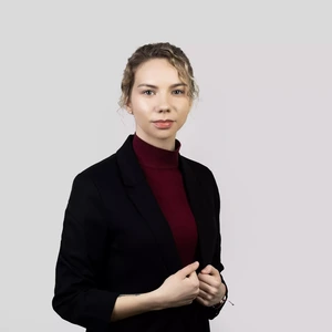 Portretinė nuotrauka – moteris su šviesiais, lengvai banguotais plaukais, surištais į kuodelį. Ji vilki tamsų švarką ir bordo spalvos golfą. Fonas – šviesiai pilkas su subtiliu rausvu atspalviu dešinėje pusėje.