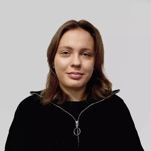 Portretinė nuotrauka – jauna moteris rudais plaukais, vilkinti juodą užsegamą megztinį su apvaliu žiediniu užtrauktuko užsegimu. Ji žiūri į kamerą neutraliame pilkame fone.