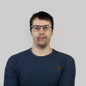 Portretinė nuotrauka – jaunas vyras tamsiais plaukais ir akiniais, vilkintis mėlyną megztinį, žvelgia tiesiai į kamerą šviesiame fone.