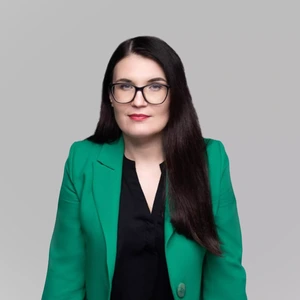 Portretinė nuotrauka – moteris ilgais tamsiais plaukais, dėvinti ryškiai žalią švarką ir juodą palaidinę. Ji žiūri tiesiai į kamerą per akinius, švelniai šypsosi. Fonas neutralus, šviesiai pilkas.
