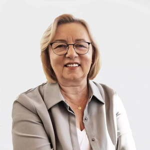 Brandesnio amžiaus moteris su šviesiais trumpais plaukais, dėvinti akinius, šviesiai pilką švarkelį ir baltus marškinėlius, šypsosi prieš šviesų foną.
