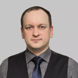 Portretinė nuotrauka – vyras trumpais tamsiais plaukais, dėvintis dryžuotus marškinius, tamsų liemenę ir blizgų mėlyną kaklaraištį. Jis rimtai žiūri į kamerą. Fonas neutralus, pilkšvas.