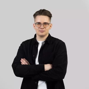 Portretinė nuotrauka – jaunas vyras trumpais šviesiai rudais plaukais, dėvintis apvalius akinius. Apsirengęs baltus marškinėlius ir juodą marškinių tipo švarkelį. Rankos sukryžiuotos. Fonas – šviesiai pilkas